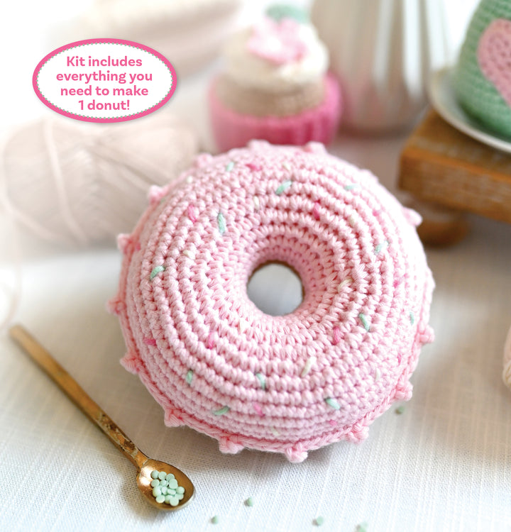 The Crochet Cafe Amigurumi Kit Donut by Schiffer Publishing, Ltd. features a pink crocheted donut with light blue sprinkles on a white surface beside a gold spoon and blurred crochet supplies in the background. Text highlights its beginner focus.