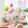 A display of Schiffer Publishing, Ltd.’s Crochet Cafe Amigurumi Kit Donut sits among pastel dishes, yarn, and a teapot. Text in a pink bubble says, "Collect the whole set!.