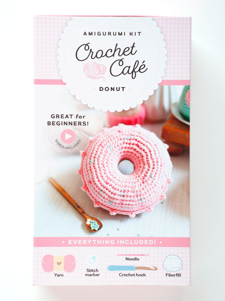 The Crochet Cafe Amigurumi Kit Donut by Schiffer Publishing, Ltd. features a pink and white crochet donut and includes yarn, stitch marker, crochet hook, needle, and fiberfill—ideal for beginner crochet enthusiasts.