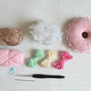 The Crochet Cafe Amigurumi Kit Donut by Schiffer Publishing, Ltd. includes brown and pink yarn, small green, yellow, and pink bundles, white stuffing, a pink donut project—great for beginners—plus a blue stitch marker, black hook, and metal needle.