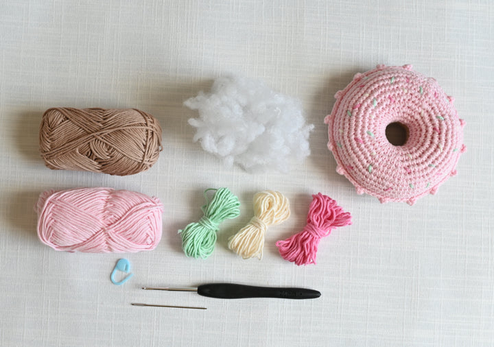The Crochet Cafe Amigurumi Kit Donut by Schiffer Publishing, Ltd. includes brown and pink yarn, small green, yellow, and pink bundles, white stuffing, a pink donut project—great for beginners—plus a blue stitch marker, black hook, and metal needle.