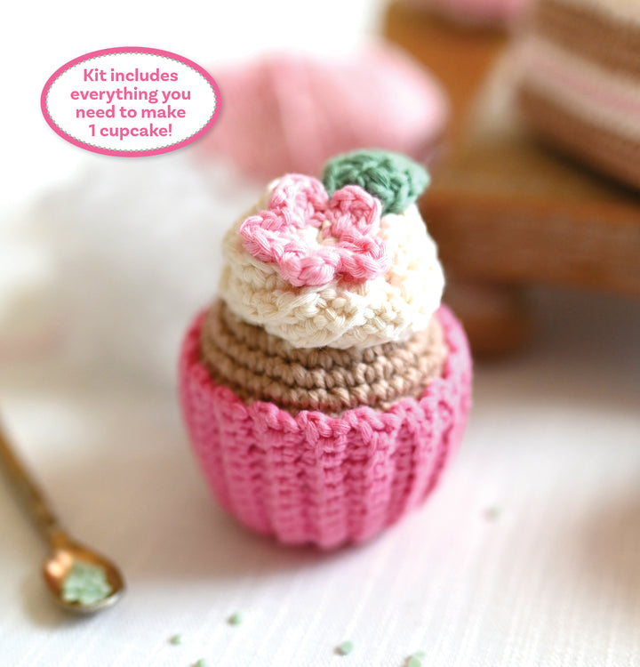 The Crochet Cafe Amigurumi Kit Cupcake by Schiffer Publishing, Ltd. includes supplies to make a cupcake with a pink base, beige middle, white top, pink flower and green leaf—perfect for beginners! Comes with a small spoon and text bubble.