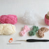 Yarn, stuffing, a crocheted cupcake, crochet hook, sewing needle, and stitch marker from the Schiffer Publishing, Ltd. Crochet Cafe Amigurumi Kit Cupcake are neatly displayed on a light surface—ideal for showcasing beginner-friendly materials.