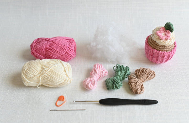 Yarn, stuffing, a crocheted cupcake, crochet hook, sewing needle, and stitch marker from the Schiffer Publishing, Ltd. Crochet Cafe Amigurumi Kit Cupcake are neatly displayed on a light surface—ideal for showcasing beginner-friendly materials.