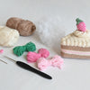 The Crochet Cafe Amigurumi Kit Cake Slice by Schiffer Publishing, Ltd. includes a crocheted cake slice with a pink strawberry, yarn balls, short bundles, and tools—perfect for making sweet amigurumi treats.