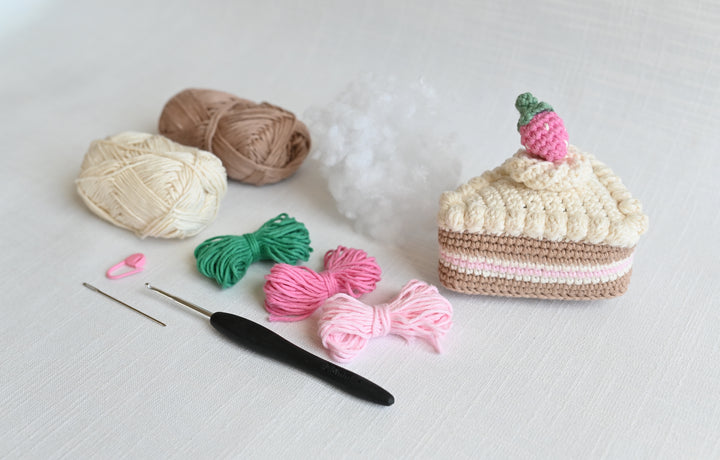 The Crochet Cafe Amigurumi Kit Cake Slice by Schiffer Publishing, Ltd. includes a crocheted cake slice with a pink strawberry, yarn balls, short bundles, and tools—perfect for making sweet amigurumi treats.