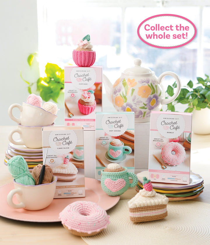 A display of Schiffer Publishing, Ltd.’s Crochet Cafe Amigurumi Kit Hot Cocoa with cupcakes, donuts, pastel yarn, stacked teacups, and a floral teapot. Text: “Collect the whole set!”.