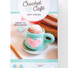 The Crochet Cafe Amigurumi Kit Hot Cocoa by Schiffer Publishing, Ltd. features a pastel box with a photo of a crochet mug topped with whipped cream and a heart, plus icons showing included yarn, needle, hook, and fiberfill.