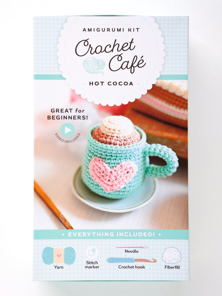 The Crochet Cafe Amigurumi Kit Hot Cocoa by Schiffer Publishing, Ltd. features a pastel box with a photo of a crochet mug topped with whipped cream and a heart, plus icons showing included yarn, needle, hook, and fiberfill.
