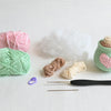 The Crochet Cafe Amigurumi Kit Hot Cocoa by Schiffer Publishing, Ltd. features yarn, stuffing, hooks, a stitch marker, and a heart mug—all on a white surface—showing ideal materials for cozy crochet or amigurumi projects.
