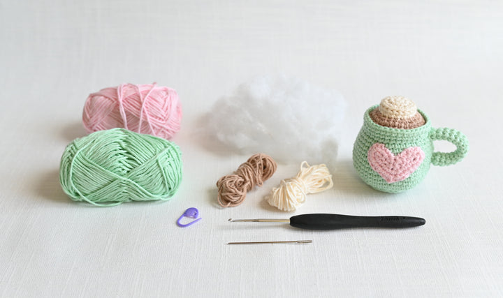 The Crochet Cafe Amigurumi Kit Hot Cocoa by Schiffer Publishing, Ltd. features yarn, stuffing, hooks, a stitch marker, and a heart mug—all on a white surface—showing ideal materials for cozy crochet or amigurumi projects.
