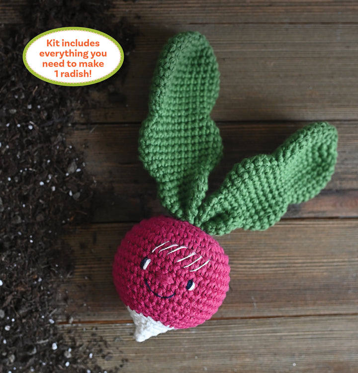 The Crochet Your Veggies Amigurumi Kit Radish by Schiffer Publishing, Ltd. features a smiling crocheted radish with two green leaves on a wooden surface beside spilled soil. The kit includes everything needed to make one radish.