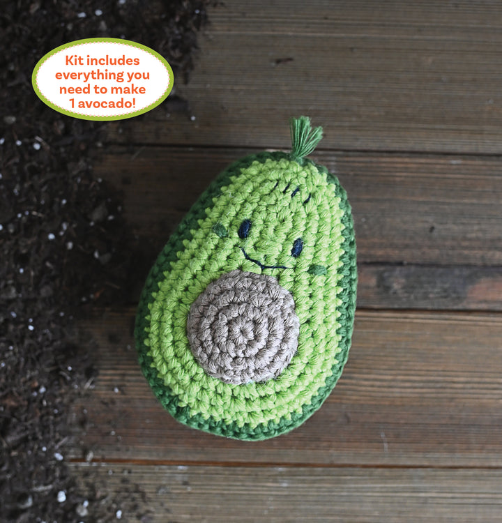 The Crochet Your Veggies Amigurumi Kit Avocado by Schiffer Publishing, Ltd. features a smiling crocheted avocado with a textured pit sitting on wood beside scattered soil. The kit includes everything you need to make 1 avocado.