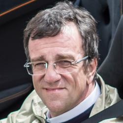 A man with short dark hair and glasses smiles subtly in a car, wearing a light jacket—reflecting the candid racing style celebrated in "Lords of Speed" by Schiffer Publishing, Ltd.