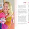 A smiling blonde woman in a colorful sweater holds bright skeins of yarn, standing in a cozy craft room filled with Handmade Happiness Yarn Crafts by Schiffer Publishing, Ltd. Shelves display vibrant yarn projects and home décor in the background.