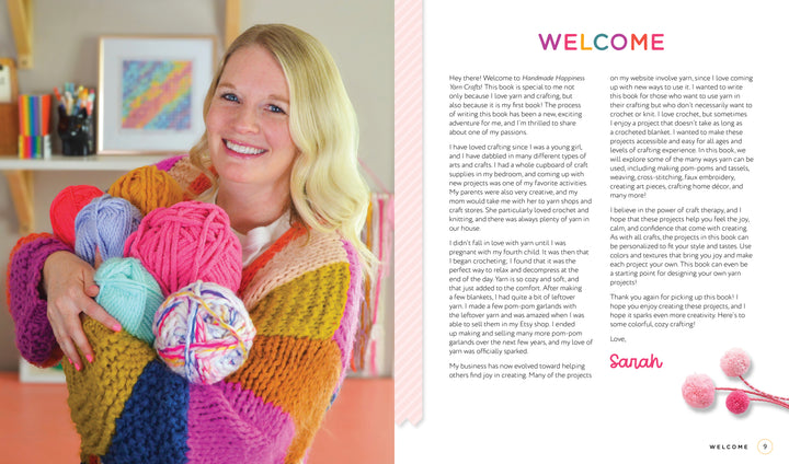 A smiling blonde woman in a colorful sweater holds bright skeins of yarn, standing in a cozy craft room filled with Handmade Happiness Yarn Crafts by Schiffer Publishing, Ltd. Shelves display vibrant yarn projects and home décor in the background.