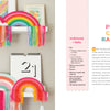 Two white shelves showcase colorful rainbow wall hangings inspired by Handmade Happiness Yarn Crafts from Schiffer Publishing, Ltd. A small cactus, calendar, and pastel artwork complete the display. Yarn crafts instructions are shown to the right.