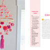 The Handmade Happiness Yarn Crafts chandelier by Schiffer Publishing, Ltd. features bright pom-poms, tassels, and wooden rings, hanging beside a white bed in a sunny room with white walls and a window.