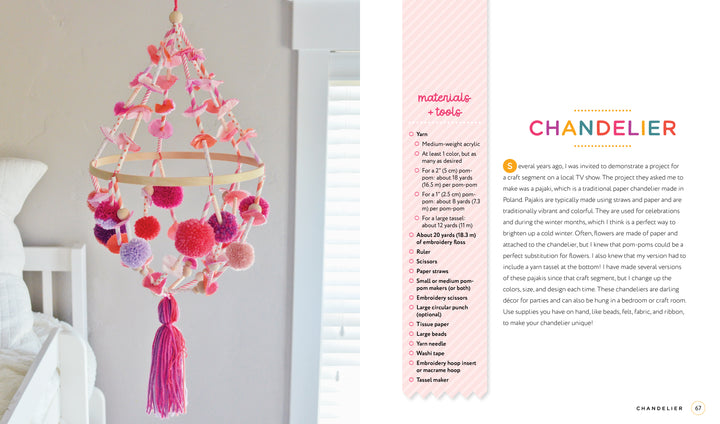 The Handmade Happiness Yarn Crafts chandelier by Schiffer Publishing, Ltd. features bright pom-poms, tassels, and wooden rings, hanging beside a white bed in a sunny room with white walls and a window.