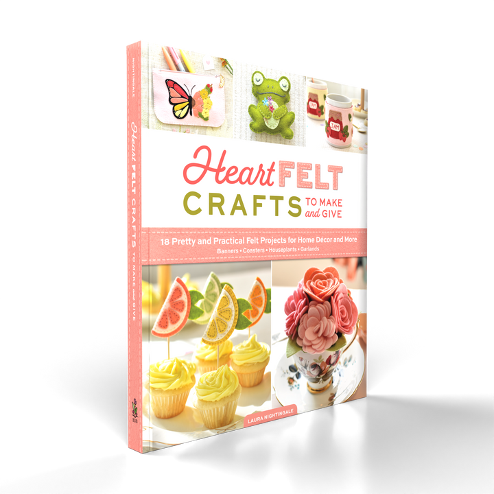 The book HeartFELT Crafts to Make and Give by Schiffer Publishing, Ltd. features colorful beginner felt projects like a frog, butterfly, fruit, and flower on its cover and stands upright against a white background.