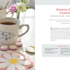 A cup of tea in a white mug with blue flowers sits on a colorful daisy-shaped felt coaster—a fun beginner project from "HeartFELT Crafts to Make and Give" by Schiffer Publishing, Ltd. More coasters, a saucer, and another mug are on the sunlit table.