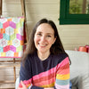 A woman with long brown hair, in a navy sweater with rainbow stripes, smiles on a chair. Behind her is a pastel patchwork quilt—perfect inspiration for beginner embroidery projects like those in "Stitching Super-Cute Food Kawaii Style" by Schiffer Publishing, Ltd.