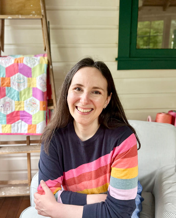 A woman with long brown hair, in a navy sweater with rainbow stripes, smiles on a chair. Behind her is a pastel patchwork quilt—perfect inspiration for beginner embroidery projects like those in "Stitching Super-Cute Food Kawaii Style" by Schiffer Publishing, Ltd.