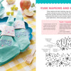 A close-up of turquoise napkins embroidered with super-cute food from "Stitching Super-Cute Food Kawaii Style" by Schiffer Publishing, Ltd., held by pastel floral napkin rings on blue plates over a yellow checkered tablecloth.
