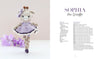 A crocheted giraffe doll in a purple dress stands on a white surface, with purple flowers to the left and Schiffer Publishing’s "Sweet and Simple Amigurumi Friends" amigurumi pattern for Sophia the Giraffe to the right.