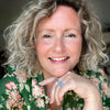 A smiling woman with curly blonde hair and a green floral shirt rests her chin on her hand, wearing a blue stone ring. She sits in soft natural light, ready to share "Yummy Watercolor" by Schiffer Publishing, Ltd. for beginners.