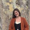 A woman with short brown hair and an orange cardigan smiles by a tree trunk—capturing a scene perfect for "The World in Me" by Schiffer Publishing, Ltd.