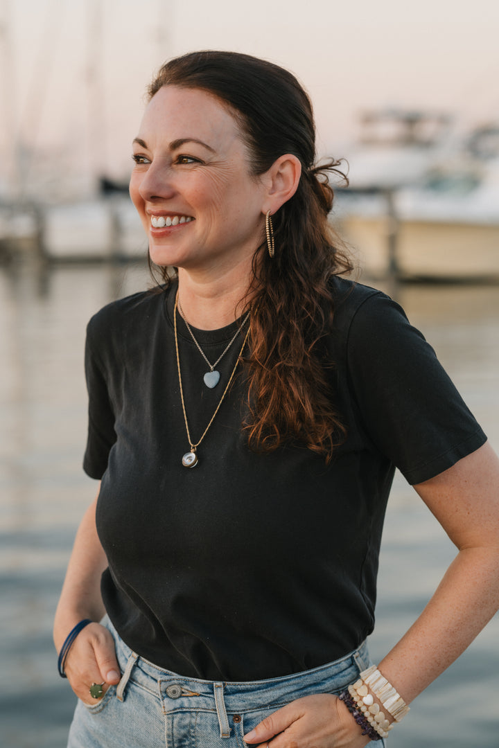 A woman with long brown hair, wearing a black t-shirt and jewelry, smiles by the marina at sunset, holding "Mermaid Candy" by Schiffer Publishing, Ltd., as if treasuring sea glass from a mermaid story.
