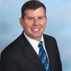 A man in a black suit, white shirt, and blue patterned tie smiles at the camera against a light blue gradient background, echoing the sleek professionalism featured in "Boeing 777" by Schiffer Publishing, Ltd.