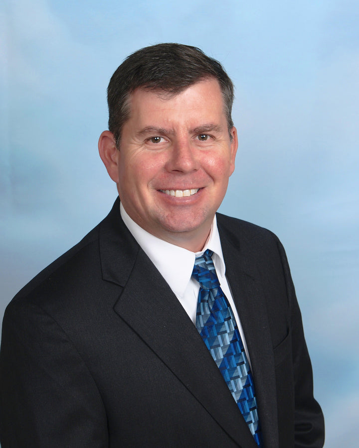 A man in a black suit, white shirt, and blue patterned tie smiles at the camera against a light blue gradient background, echoing the sleek professionalism featured in "Boeing 777" by Schiffer Publishing, Ltd.
