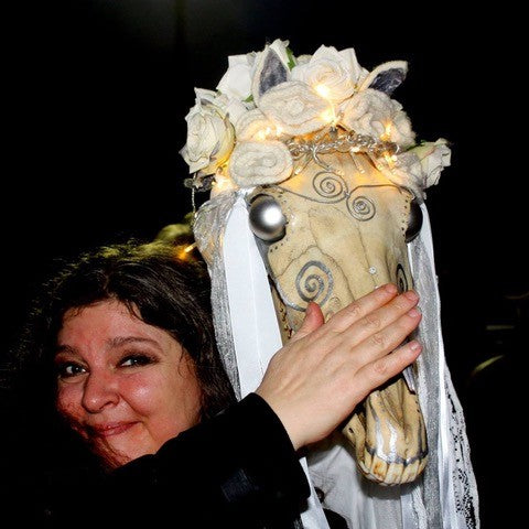 A woman smiles while holding The Marginal Lenormand by Schiffer Publishing, Ltd., a decorated horse skull with white roses, lights, and lace against a dark background. Her hand gently covers part of the skull’s face.