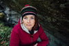 A woman with blue eyes and red braided hair sits outdoors by a large rock, wearing a red jacket and knit hat, appearing immersed in Nordic mystery as she uses the Nordic Forest Rituals Oracle Cards from Schiffer Publishing, Ltd.