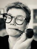 A person with short hair and round glasses smiles slightly while holding a pipe, evoking the quiet mystery of forest spirits—an atmosphere captured by Schiffer Publishing, Ltd.'s Nordic Forest Rituals Oracle Cards.