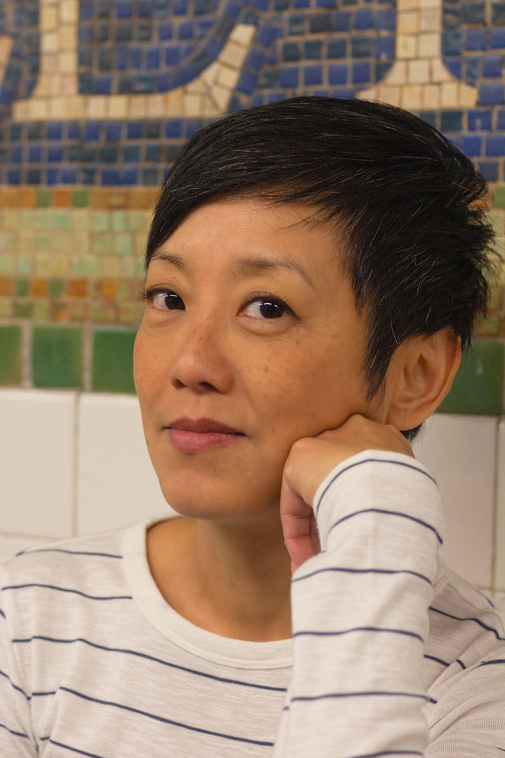 A person with short dark hair wearing a white shirt with blue stripes sits before a mosaic wall inspired by New York MTA Arts & Design, evoking the style featured in "Museum in Transit" by Schiffer Publishing, Ltd.