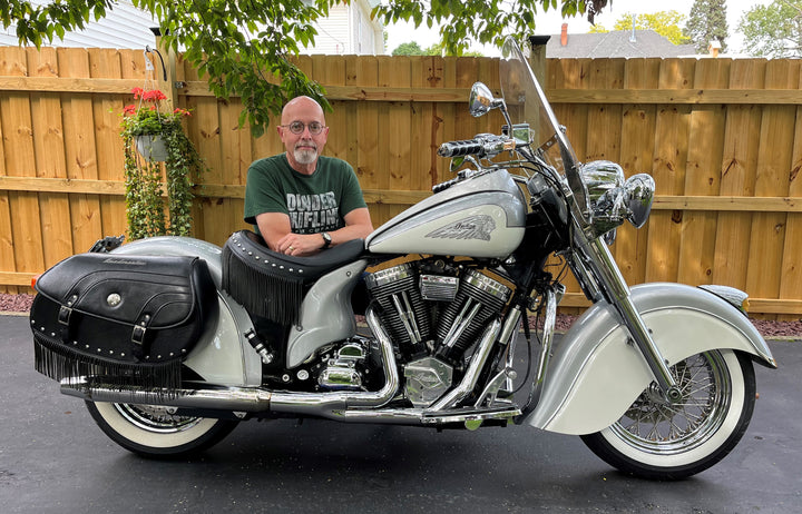 A man with glasses and a beard sits on a Gilroy Indians motorcycle by Schiffer Publishing, Ltd., parked in a driveway with a wooden fence, plants, and trees in the background.