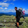 A man in hiking gear with an American flag backpack stands atop a rocky summit, green hills and distant mountains behind him. Perfect for day hikes or as inspiration from "Iconic and Hidden Gems of the Appalachian Trail" by Schiffer Publishing, Ltd.