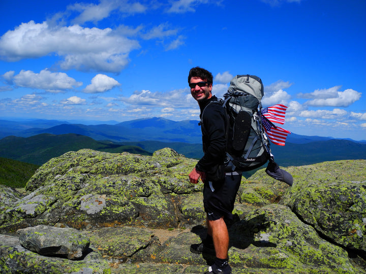 A man in hiking gear with an American flag backpack stands atop a rocky summit, green hills and distant mountains behind him. Perfect for day hikes or as inspiration from "Iconic and Hidden Gems of the Appalachian Trail" by Schiffer Publishing, Ltd.