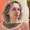 A person with curly light blue hair, glasses, gold chain earrings, and nose adornments poses in a pink sweater and striped shirt against a red-and-white backdrop reminiscent of Schiffer Publishing’s "Trees of the World.
