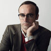 A serious-looking man in glasses and a brown blazer sits thoughtfully against a light background, appearing to ponder bird identification with "Backyard Birds of North America" by Schiffer Publishing, Ltd.