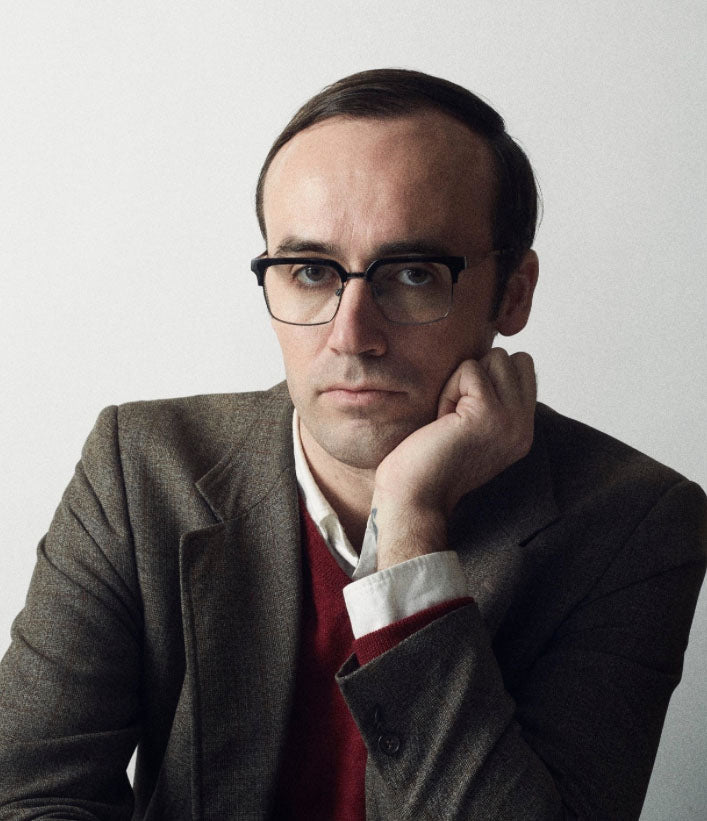 A serious-looking man in glasses and a brown blazer sits thoughtfully against a light background, appearing to ponder bird identification with "Backyard Birds of North America" by Schiffer Publishing, Ltd.