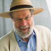 An older man with a gray beard and glasses smiles at the camera, wearing a straw hat and cream jacket. Behind Peter Laufer is a blurred background, as seen on the cover of "We Are All Americans" by Schiffer Publishing, Ltd.