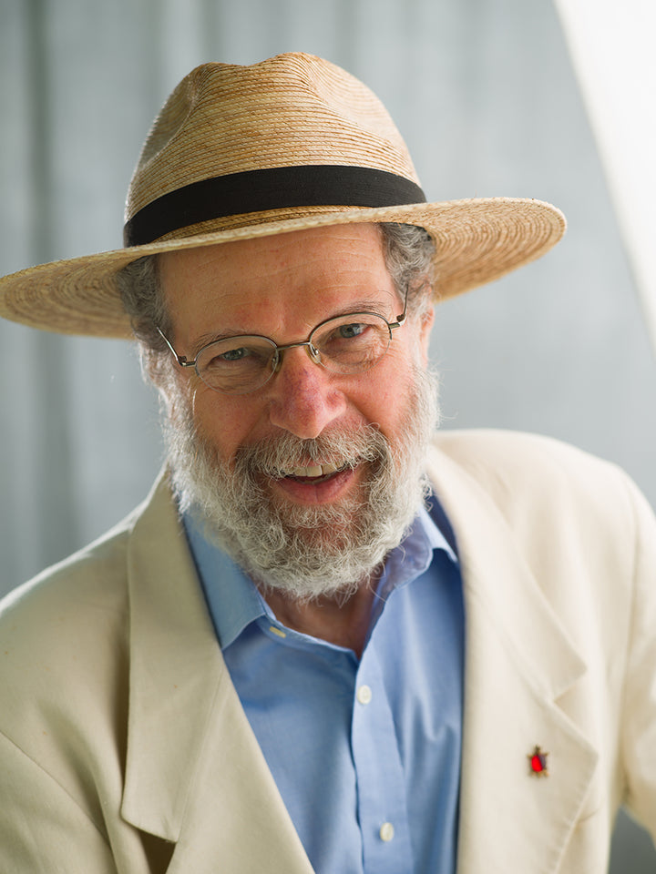 An older man with a gray beard and glasses smiles at the camera, wearing a straw hat and cream jacket. Behind Peter Laufer is a blurred background, as seen on the cover of "We Are All Americans" by Schiffer Publishing, Ltd.