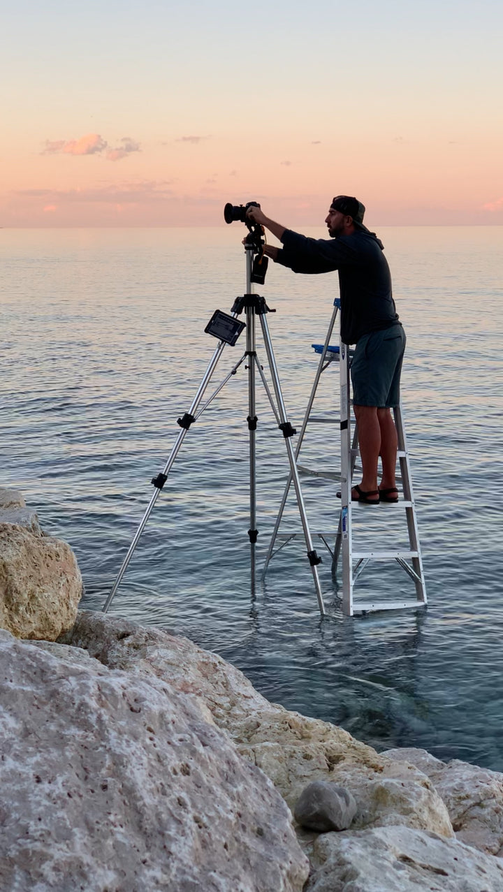 A man on a step ladder in shallow ocean water adjusts a camera at sunset, with rocks nearby and the calm sea under a pink sky—capturing scenes for Midwest Home by Schiffer Publishing, Ltd.