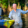 A man in a light gray shirt stands knee-deep in the lush Louisiana bayou, photographing the swamp with a tripod near his colorful kayak—an adventure inspired by "Into the Lost World" from Schiffer Publishing, Ltd.