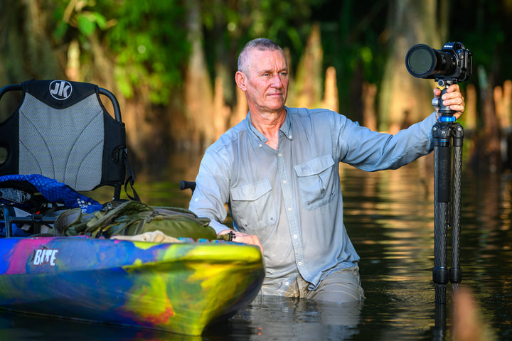 A man in a light gray shirt stands knee-deep in the lush Louisiana bayou, photographing the swamp with a tripod near his colorful kayak—an adventure inspired by "Into the Lost World" from Schiffer Publishing, Ltd.