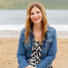 A woman with long light brown hair in a denim jacket and patterned dress smiles on a sandy beach with ocean views, enjoying seashell identification from *Seashells of Delmarva* by Schiffer Publishing, Ltd.